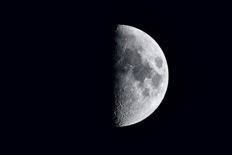 A half-lit moon on a black sky, with the right side illuminated. There are many small craters and big dark areas on the right side.