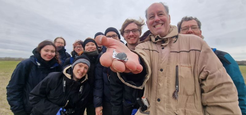 A group of young and old people in coats, standing around smiling. One man has a tinfoil square with a rock on it.