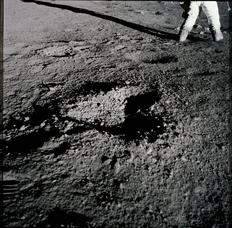 Rocky gray ground with a shallow hole with the shape of a heart, and there are space-suited legs at the top of the image.