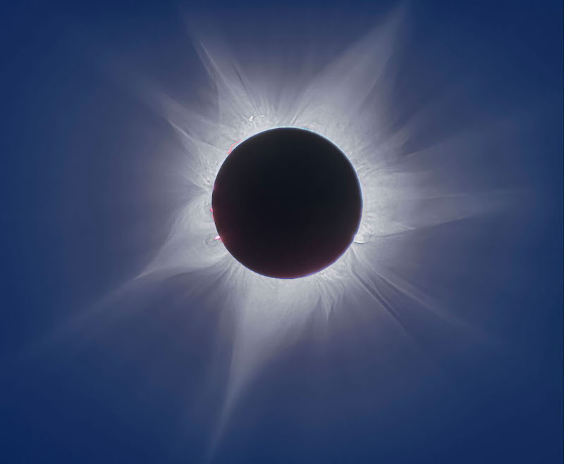 Photograph a solar eclipse: Black circle over the sun with bright white streamers showing corona and some pink promeninces along the limb.