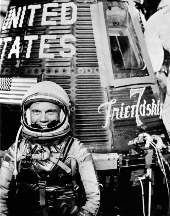 John Glenn: Black and white photo of a smiling man in space suit standing next to a conical one-person space capsule.