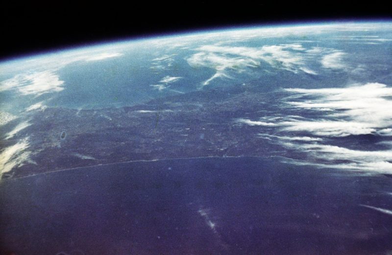 Orbital view of Earth taken by John Glenn, mostly blue sea with some white clouds, some darker land.