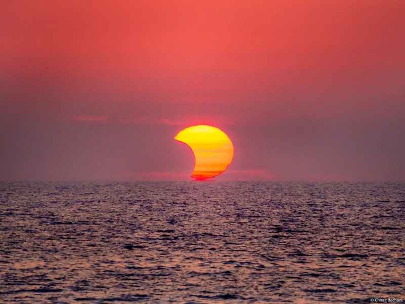 Lake with crescent sun ranging in color from red to yellow against deep orange sky.