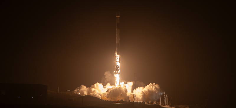 Starlink: A black and white rocket, on a pillar of golden flames, launches upward into a black night sky while clouds billow below.